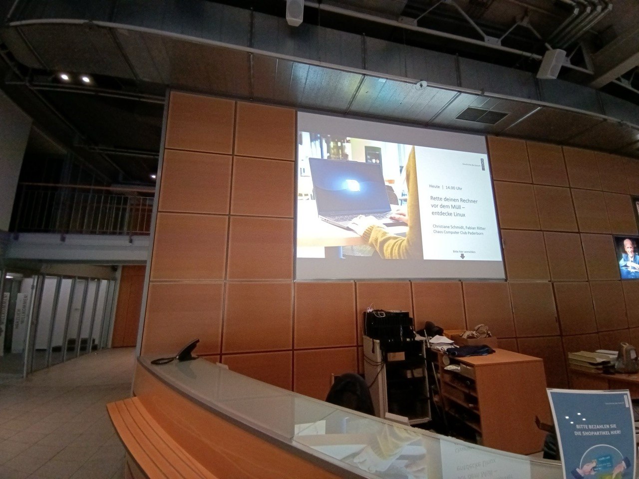 Foto vom Foyer des Heinz Nixdorf MuseumsForums, zu sehen ist der Empfangstresen, hinter dem Tresen an der Wand ist die Ankündigung der Veranstaltung auf die Wand projiziert. Links ist der Eingangsbereich Richtung Veranstaltungsräume zu sehen.