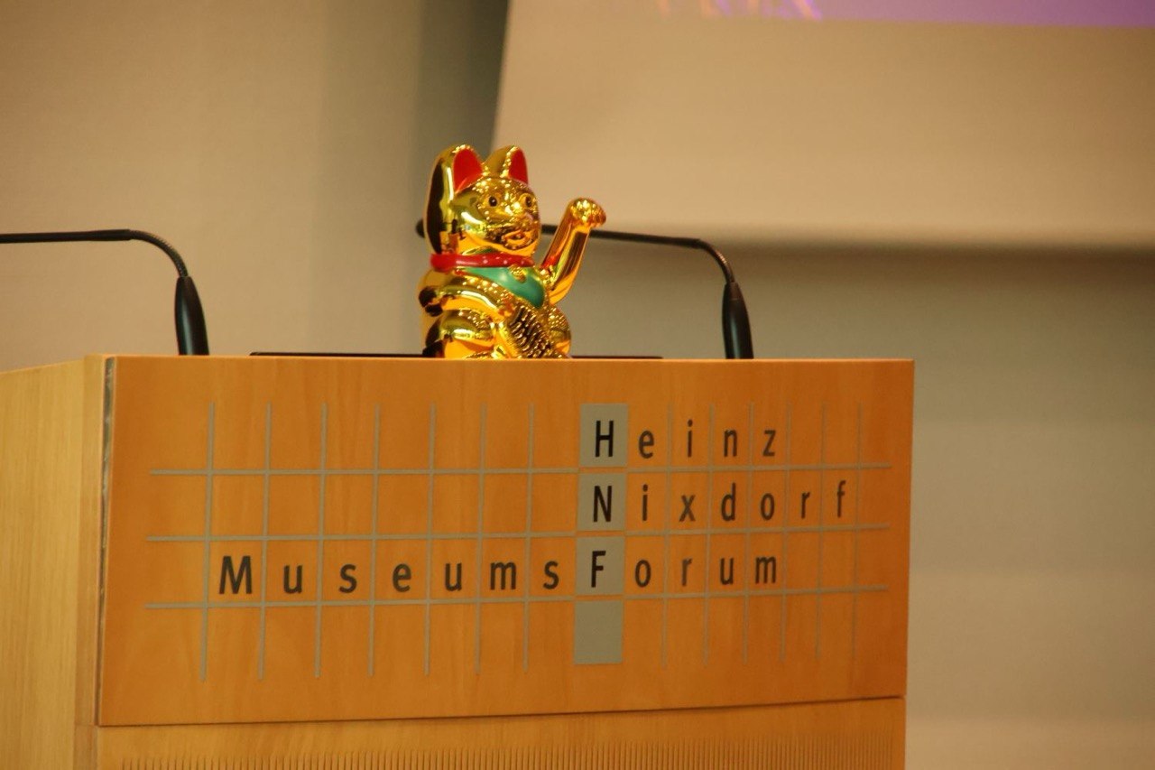 Foto eines unserer Vortragenden. Links im Bild ist eine Lichtinstallation des Subraums zu sehen. Der Vortragende steht hinter dem Rednerpult aus Holz. Vorne auf dem Pult ist der Schriftzug des Heinz Nixdorf MuseumsForums zu sehen. Oben auf dem Pult steht eine goldene Winkekatze.